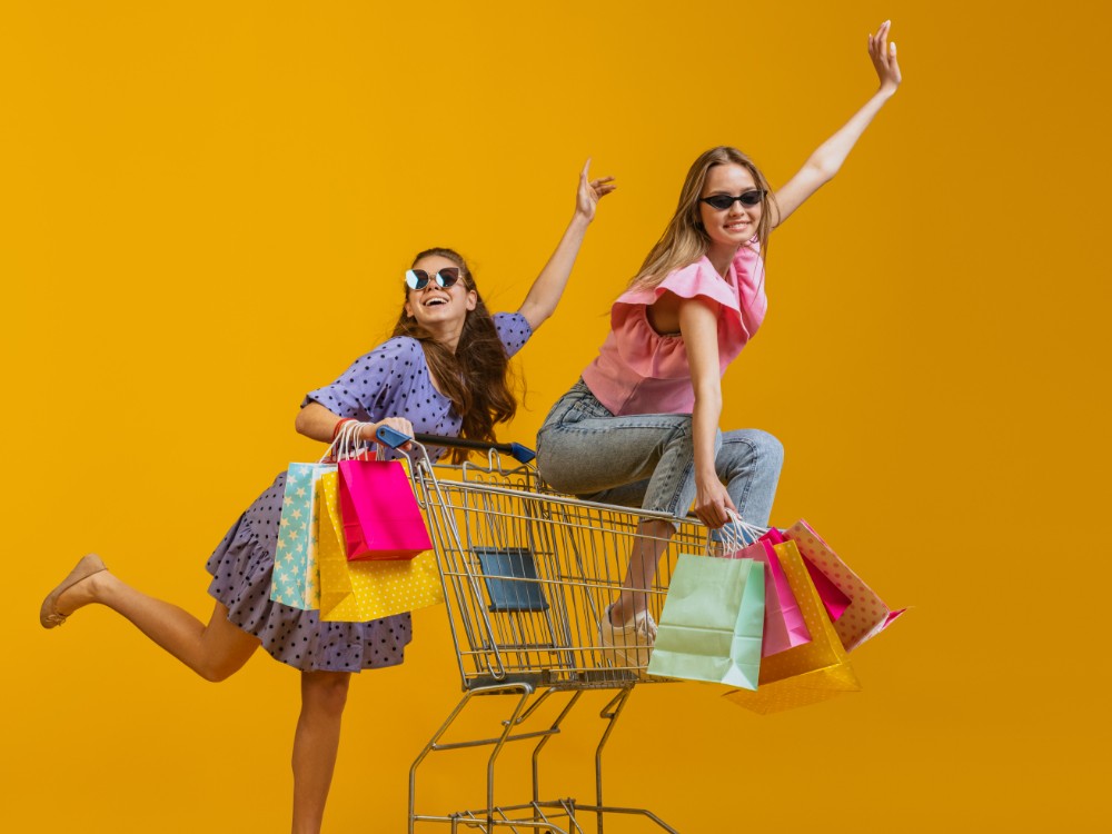 Cheerful Happy Girls With Many Shopping Bags Sitting Shopping Cart Isolated Yellow Studio Background